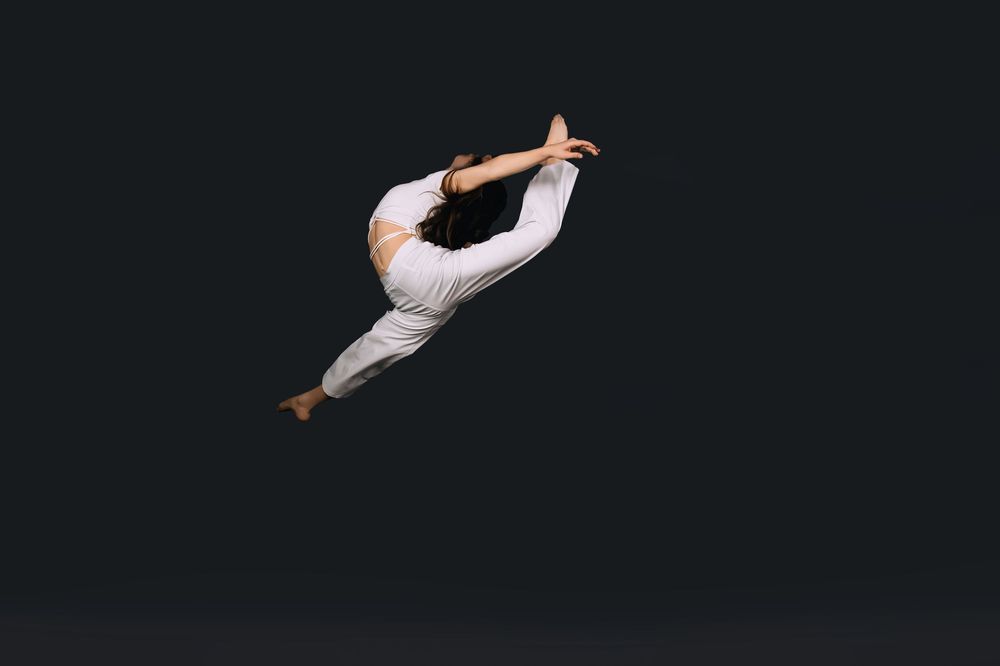 A flexible dancer leaps gracefully in white outfit against dark background, performing a stunning aerial split jump.