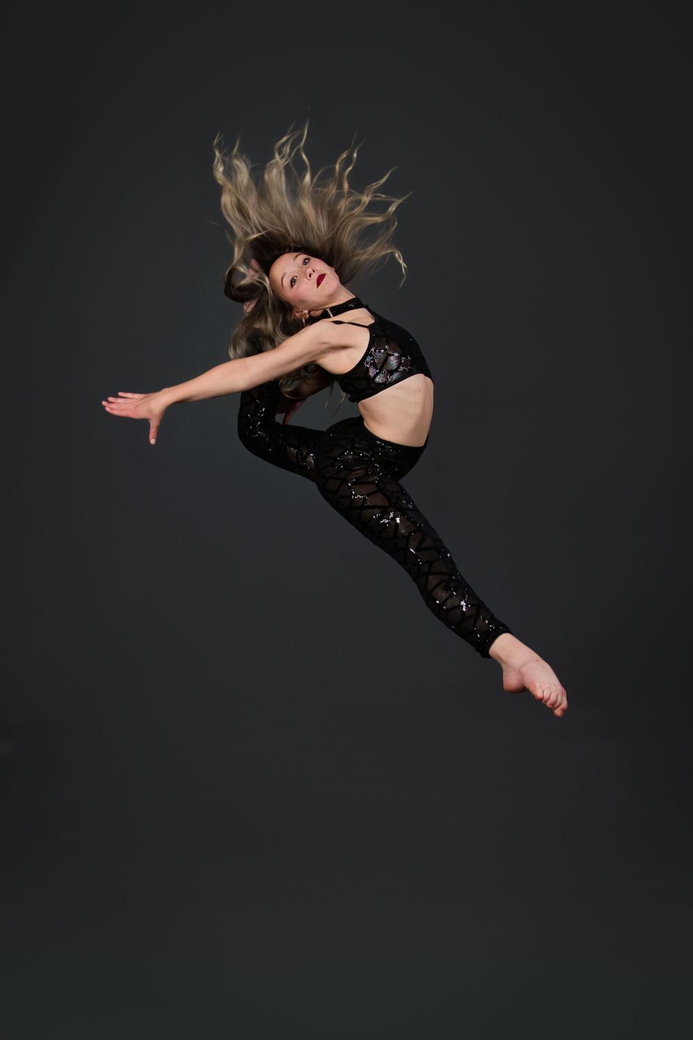 A female dancer leaps gracefully in black sequined outfit, hair flowing, against dark gray studio background.