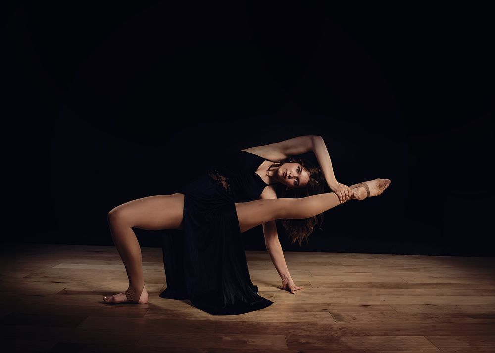 A flexible dancer in a black dress performs a dramatic floor pose on a wooden stage against a dark background.