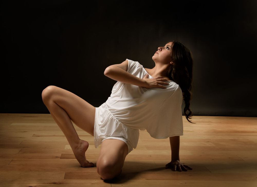 A woman in a white dress poses gracefully on a wooden floor against a dark background.