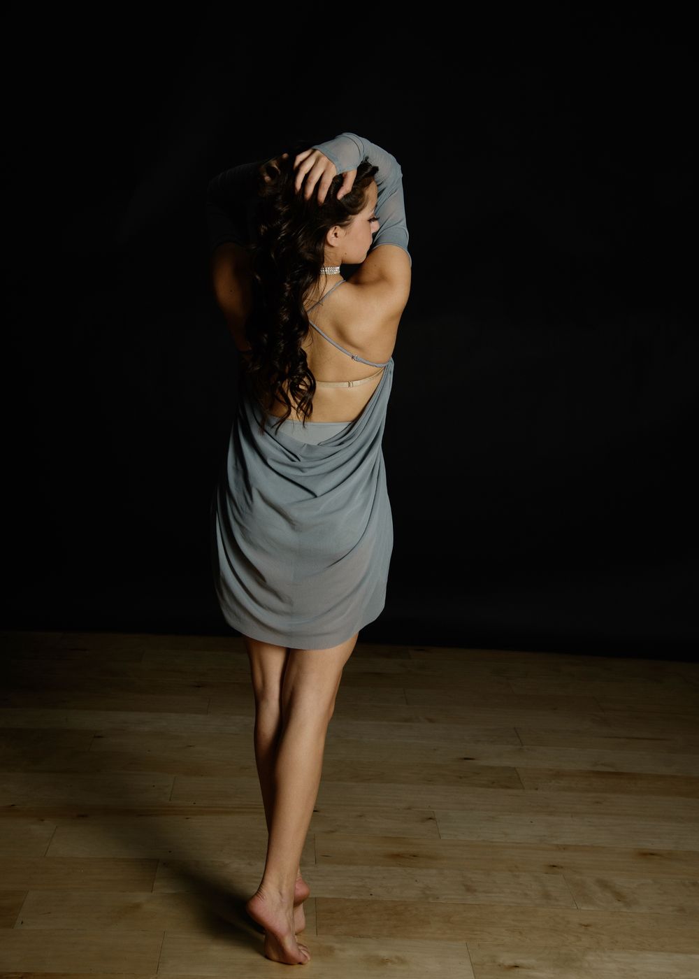 Woman in gray backless dress poses barefoot on wood floor against dark background.