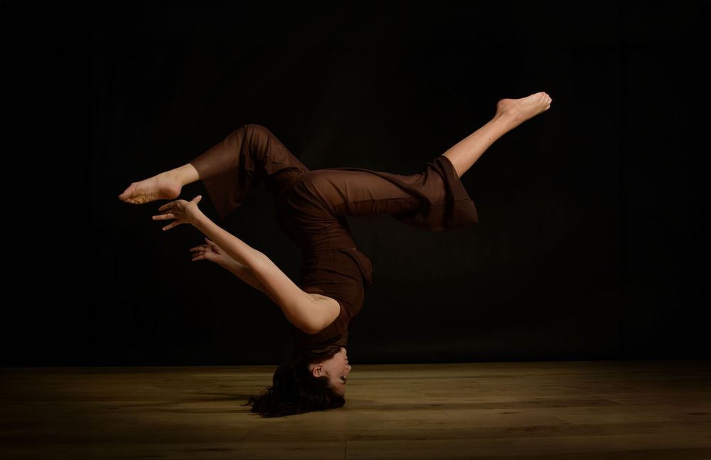 Dancer in brown outfit performs advanced headstand scorpion pose on wooden floor against dark background.