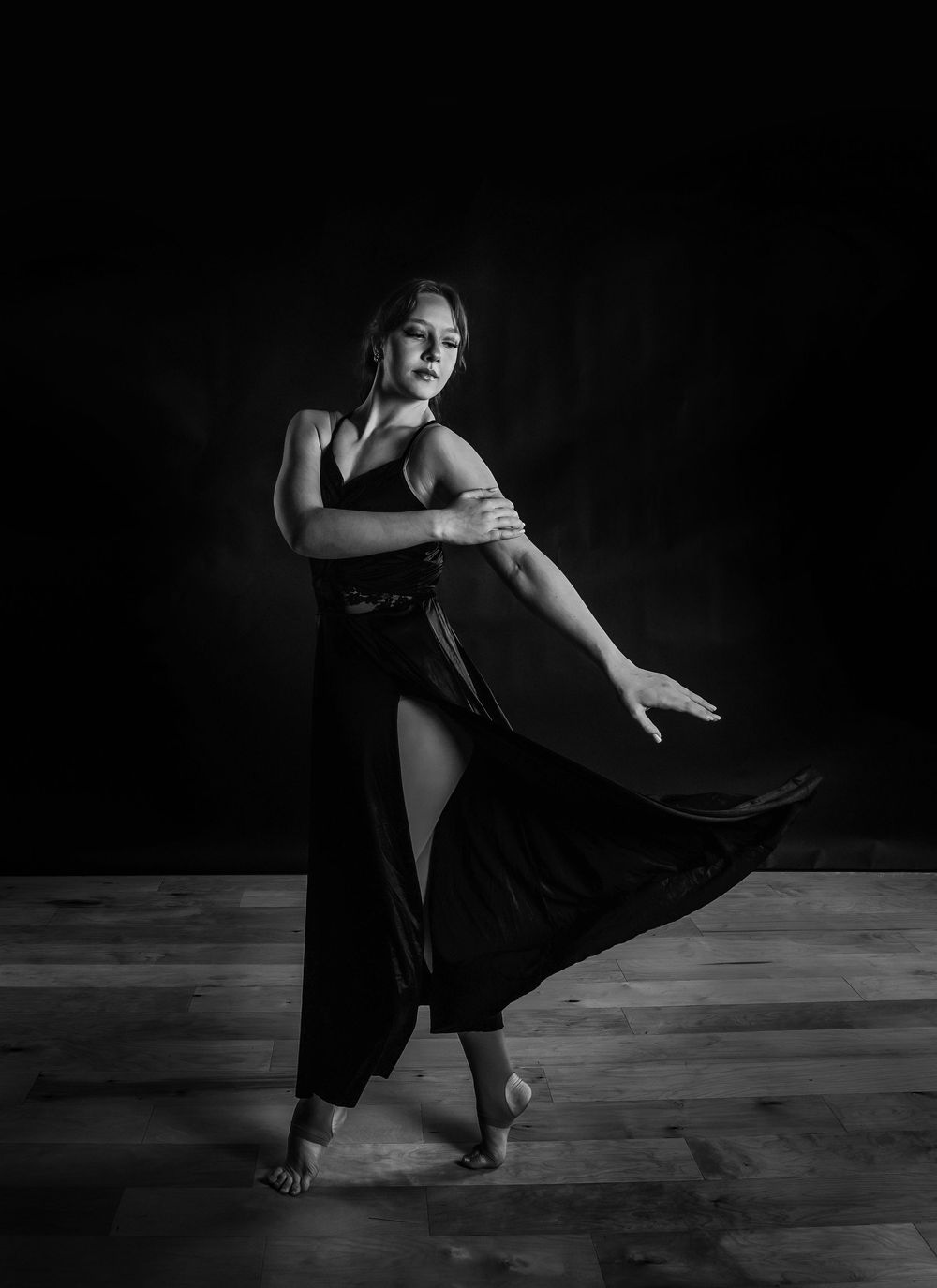 Black and white photo of a woman in a flowing black dress dancing gracefully on a wooden floor.