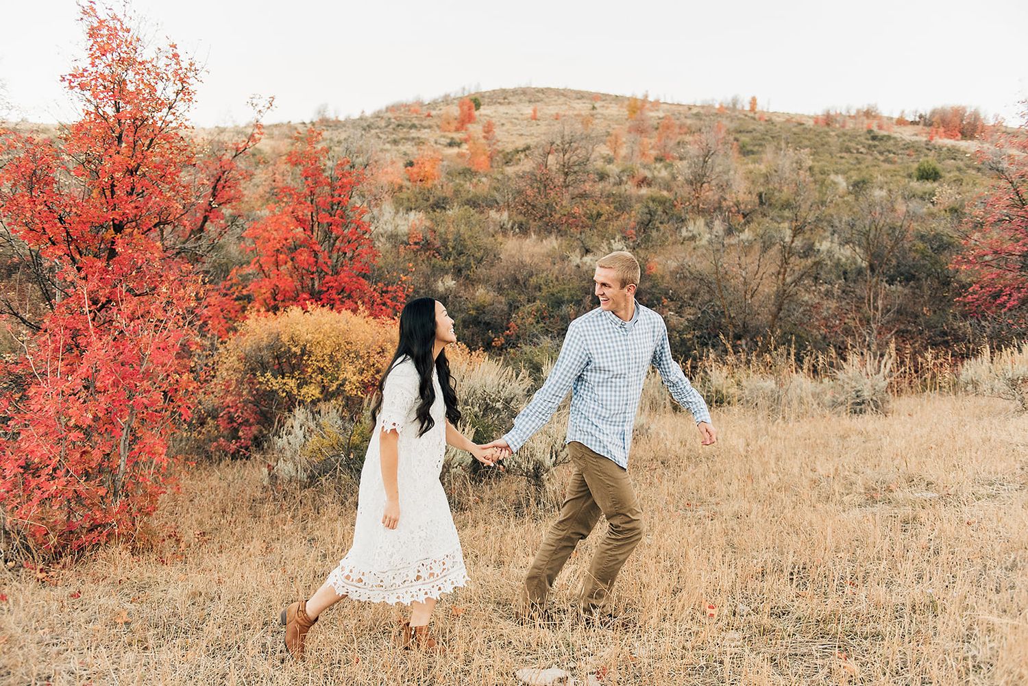 A couple holds hands while walking through autumn foliage on a hillside.