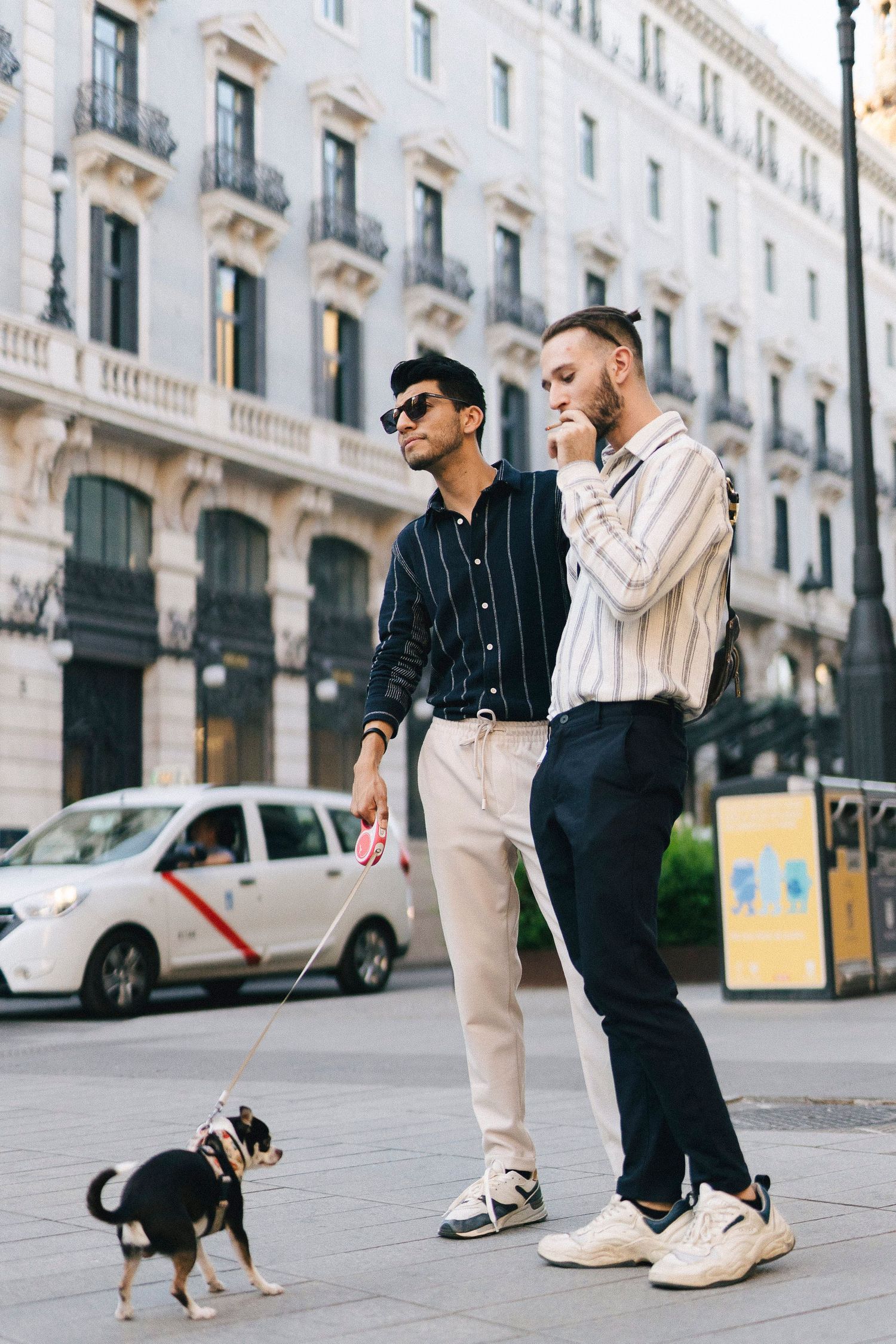 Two stylishly dressed individuals walking a small black and tan dog on a city street with classic architecture.