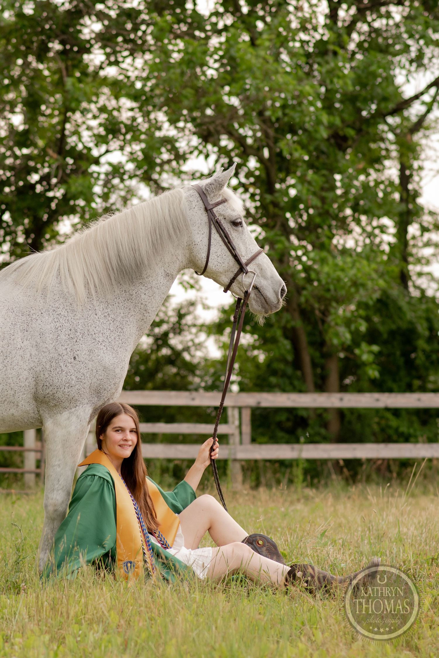 Graduate in emerald robes sits casually in grass while holding reins of a majestic white horse.