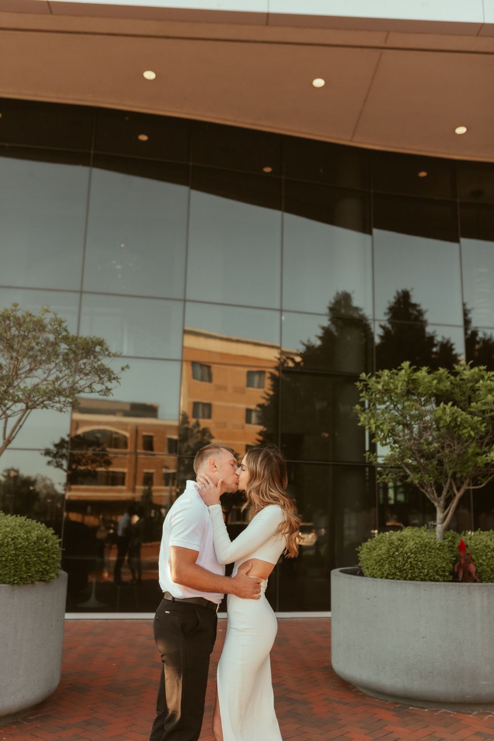 A romantic couple shares a tender moment outside a modern glass building during golden hour.