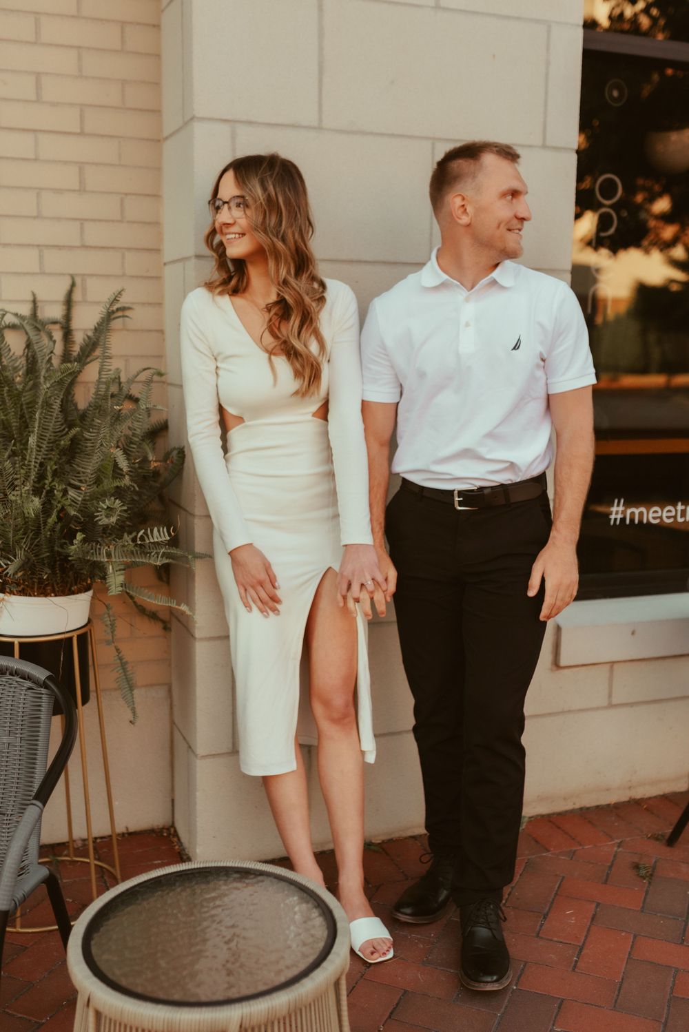 Couple in coordinated white outfits posing against brick wall with potted plants.