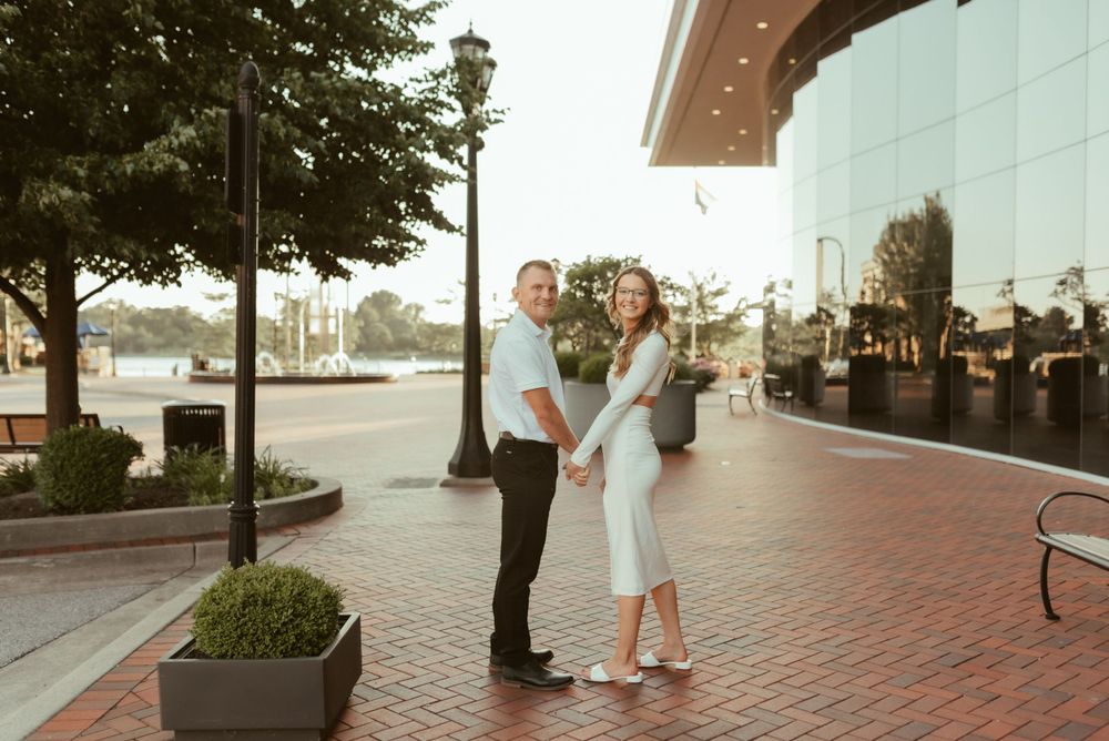 A couple poses together on a brick sidewalk at sunset near a glass building.