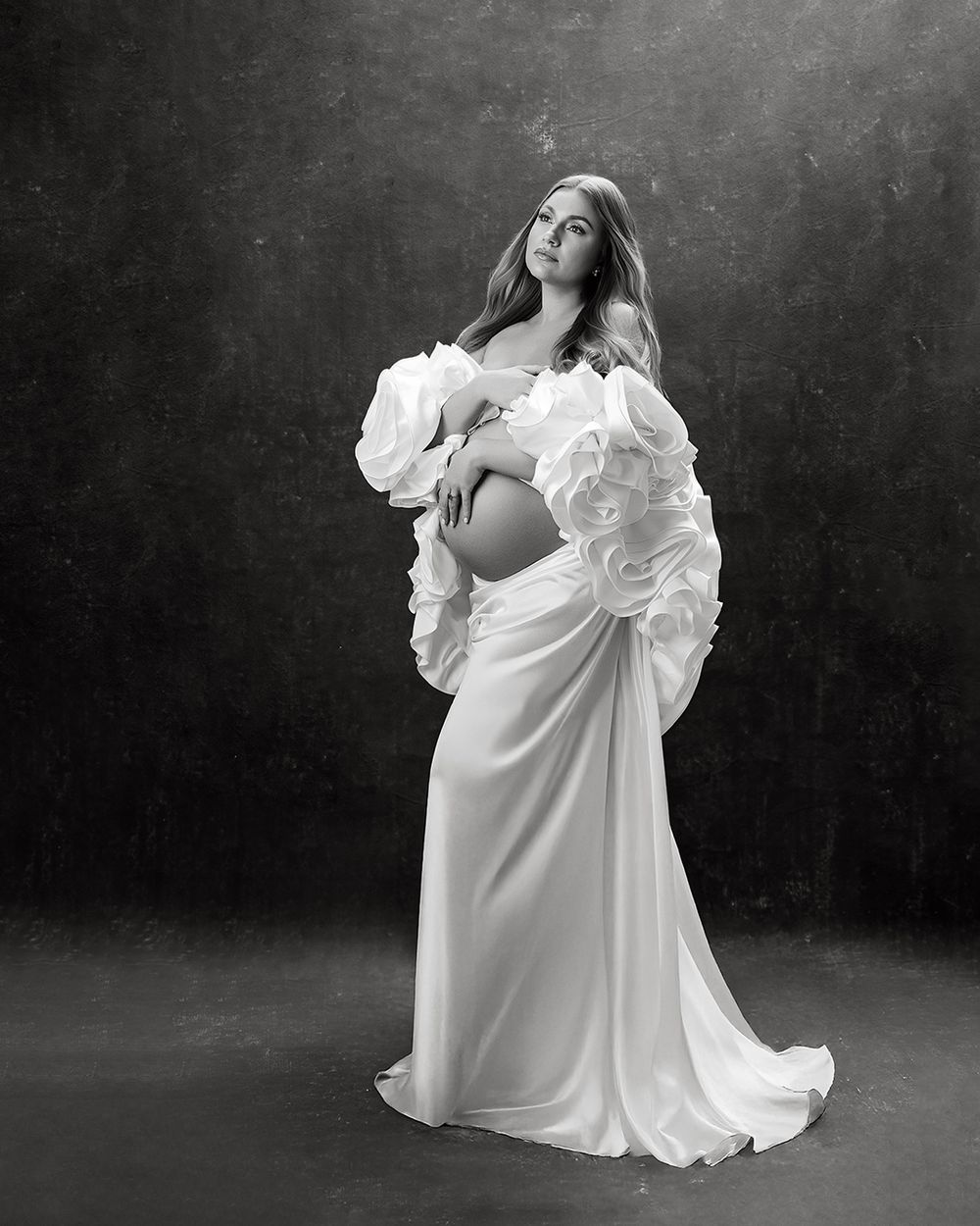 A black and white artistic maternity portrait in a  white dress with flower cape posing against a dark backdrop in Atlanta photo studio.