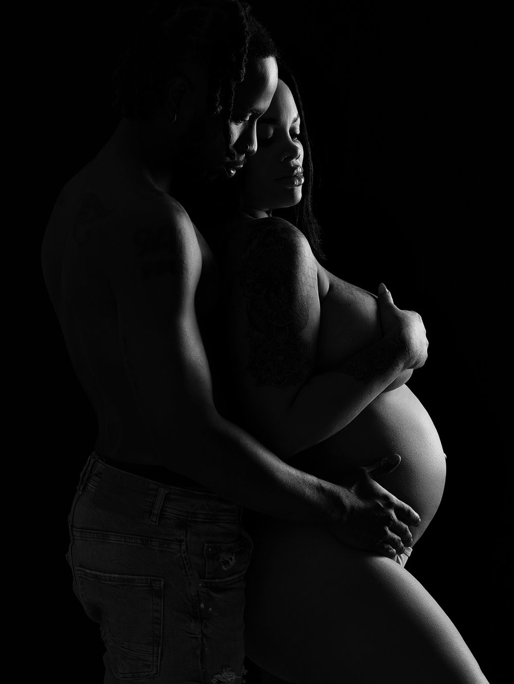 Intimate black and white maternity photo showing silhouette of couple embracing pregnant belly in dramatic lighting.