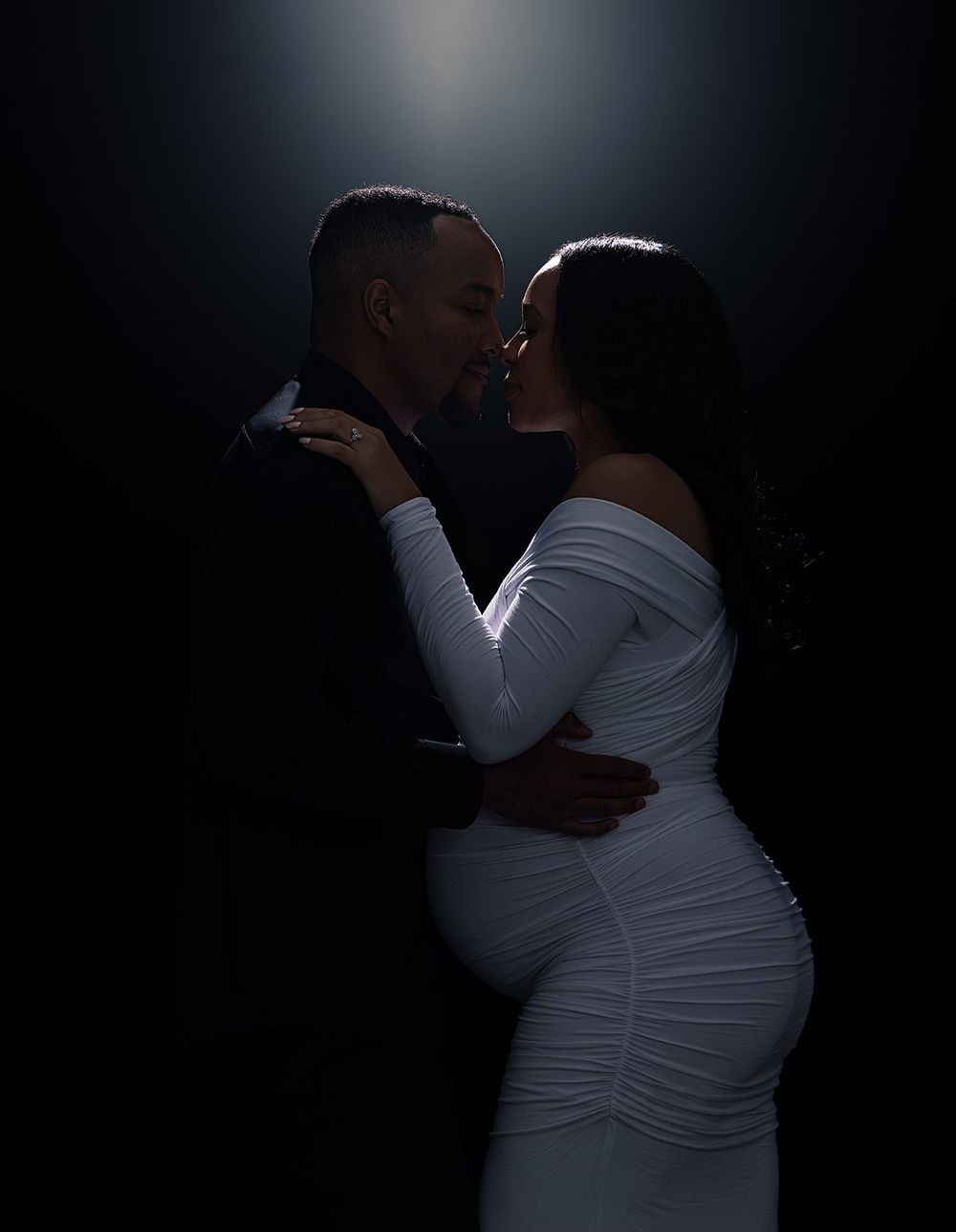 A romantic maternity photo in dim lighting shows an embracing couple in formal attire against a dark background.