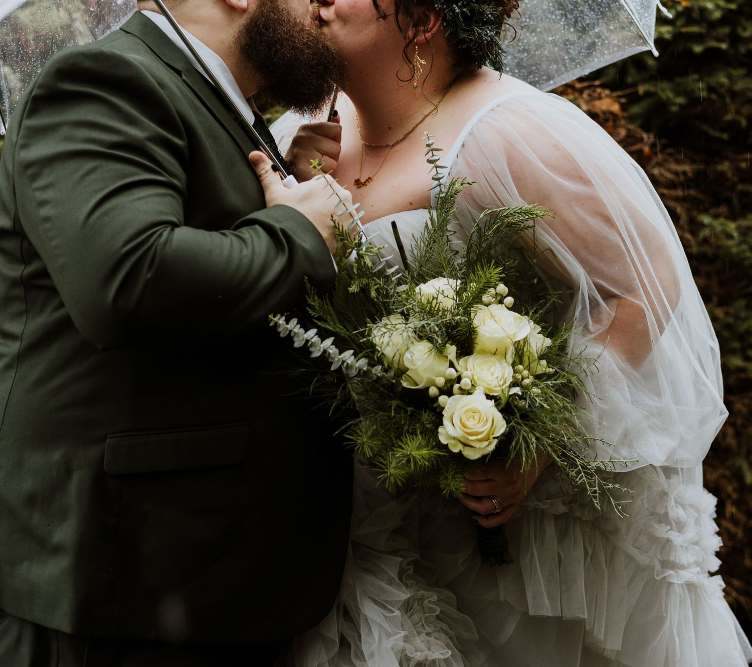 A groom in a evergreen suit kisses his bride under a clear umbrella in the rain during a Seattle Washington wedding