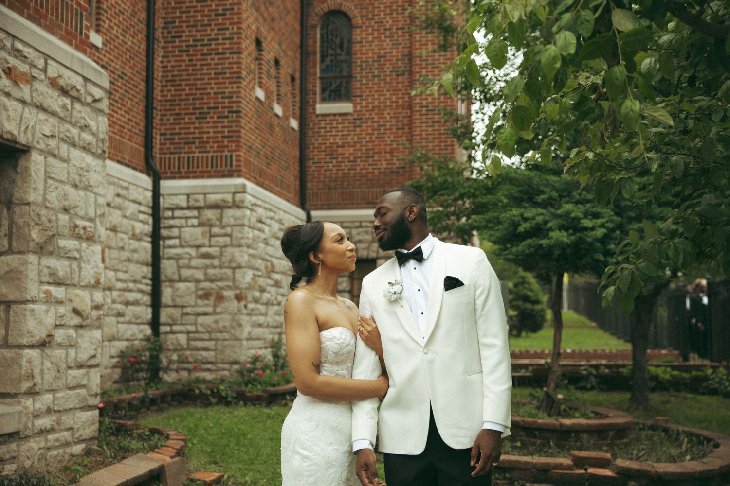 Newlyweds walking together on garden path with white gazebo in background.