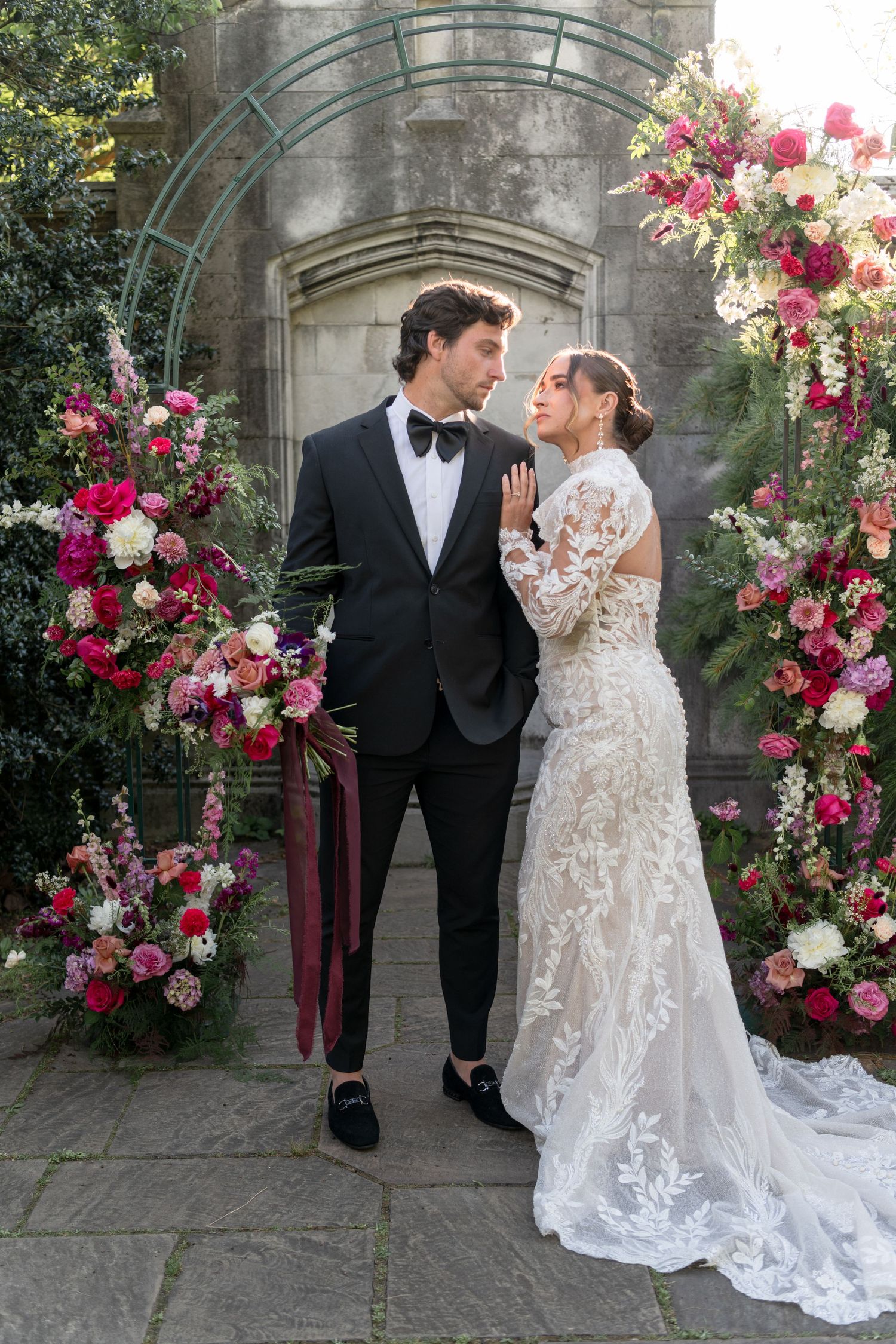Elegant couple poses under floral arch with pink and white roses in formal garden setting.