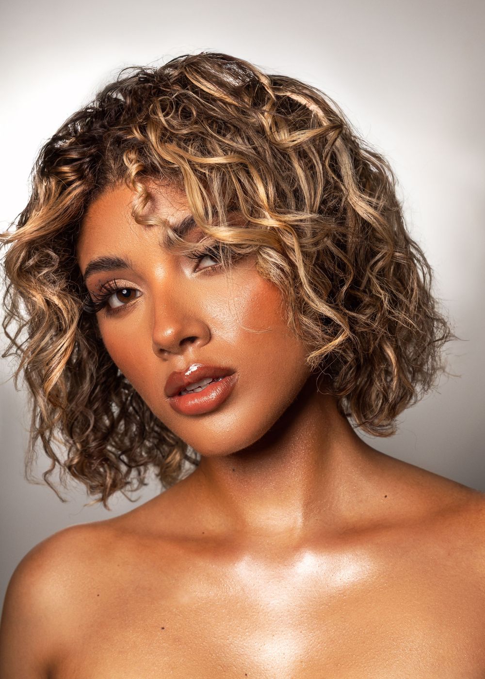 A glamour beauty portrait with highlighted curly blonde hair and glowing makeup against a light background.