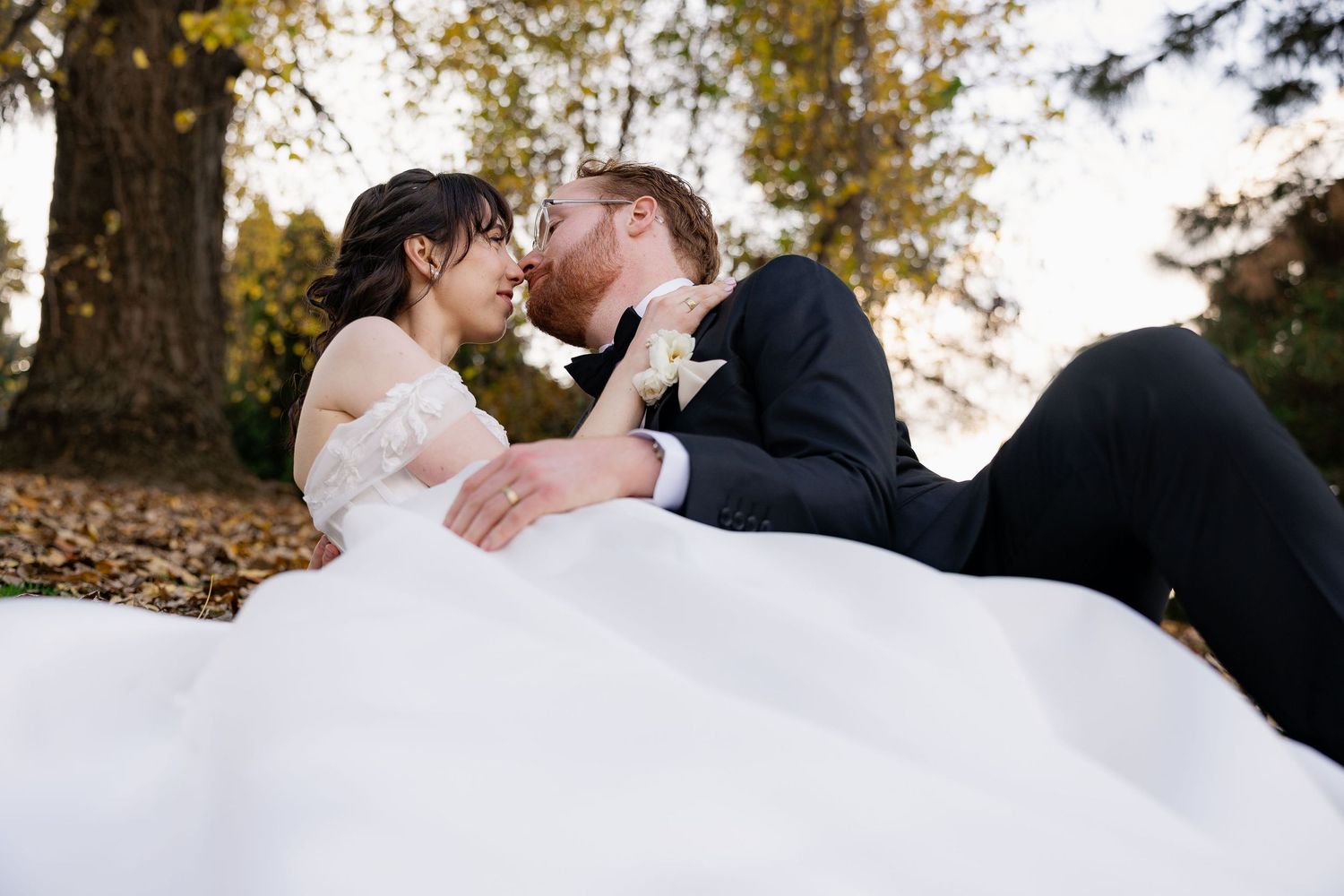 A romantic wedding couple shares a tender moment while lying on autumn leaves in an outdoor setting.