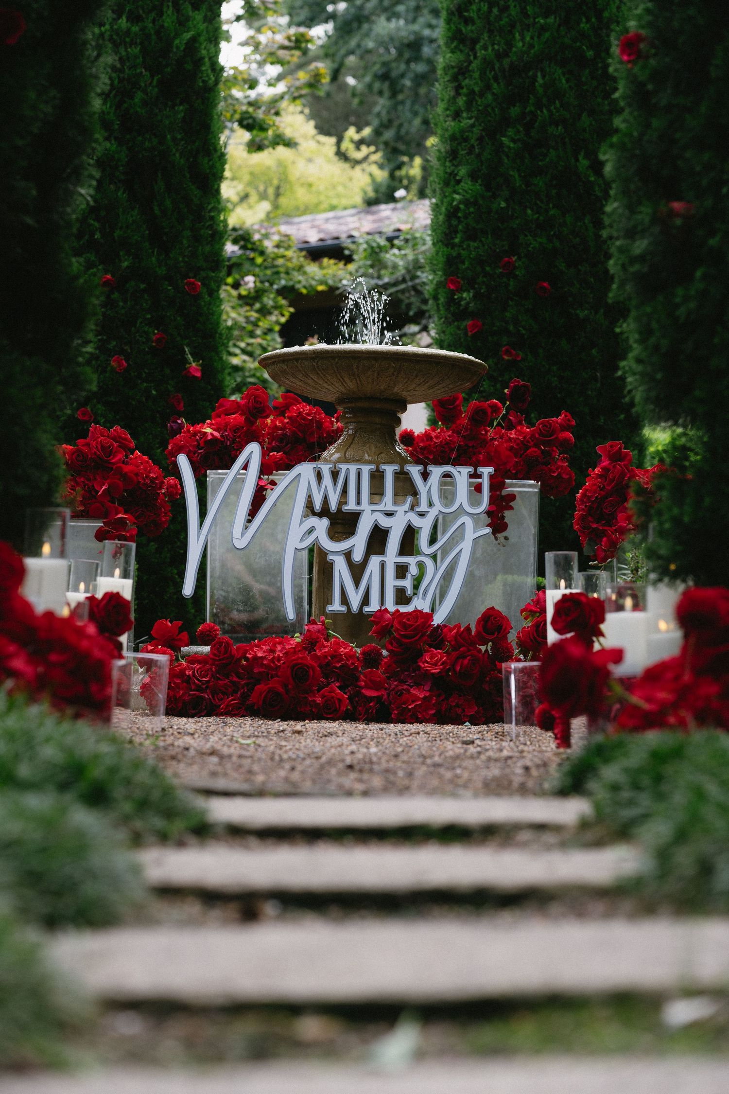 Will You Marry Me sign surrounded by red roses and fountain in garden setting.