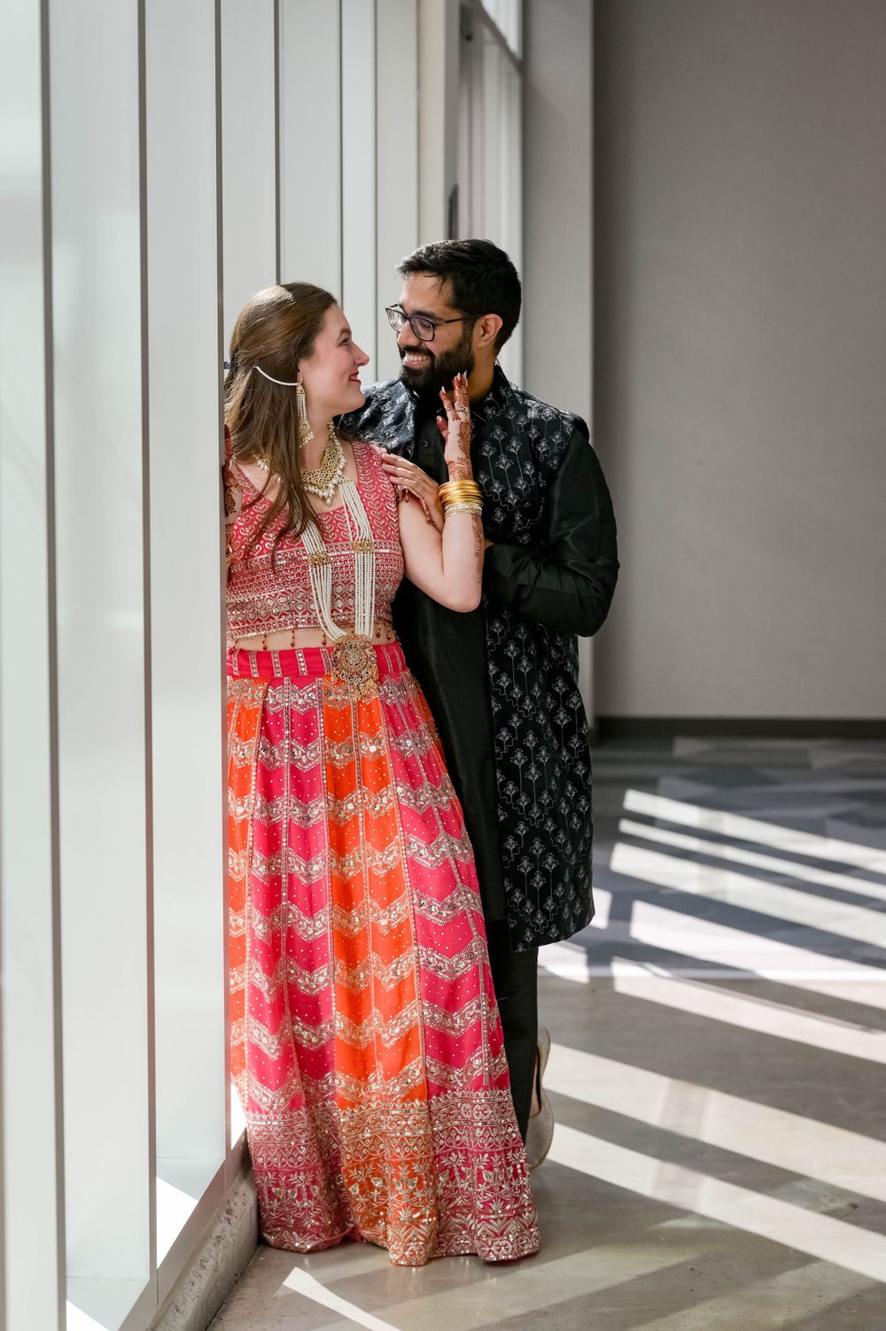 Couple in traditional Indian wedding outfits share a tender moment by large windows.