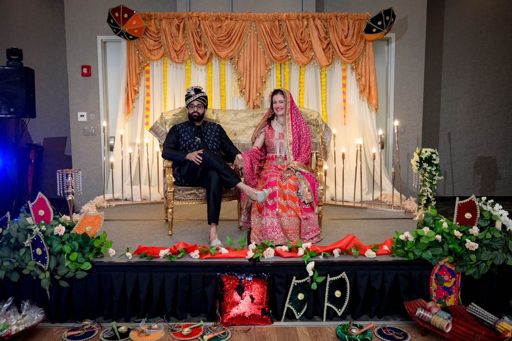 Traditional Indian wedding ceremony with couple seated on decorated stage with orange curtains and floral arrangements.