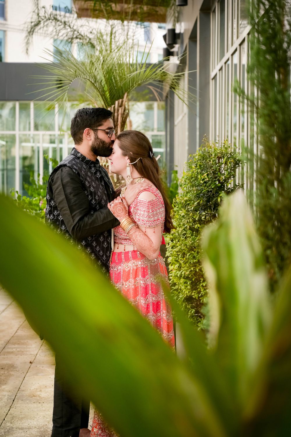 Couple in traditional Indian attire embrace among tropical plants in greenhouse setting.