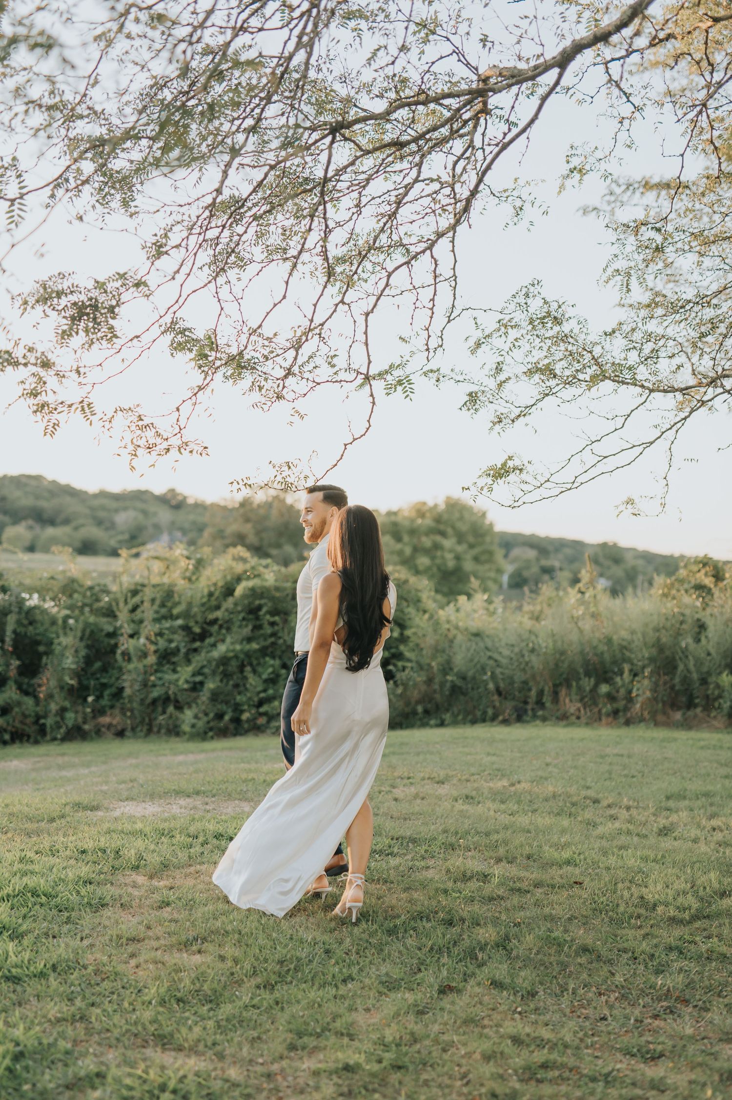 A romantic summer scene with a flowing white dress against a lush green hillside under blooming tree branches.