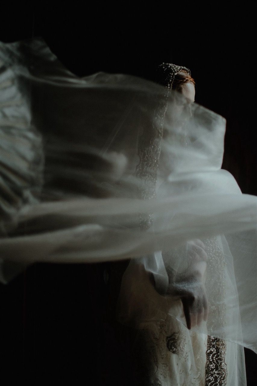 A bride in a white lace gown and headpiece with flowing veil caught in motion against a dark background.