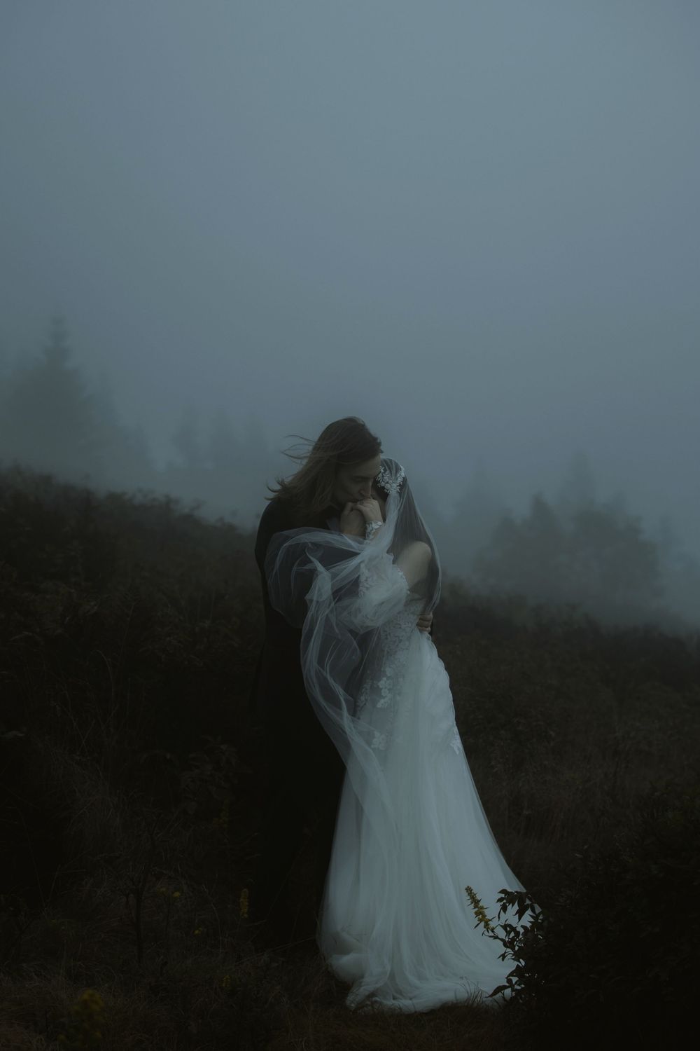 A bride and groom embrace in a misty, moody outdoor setting, her white gown and veil flowing in the fog.