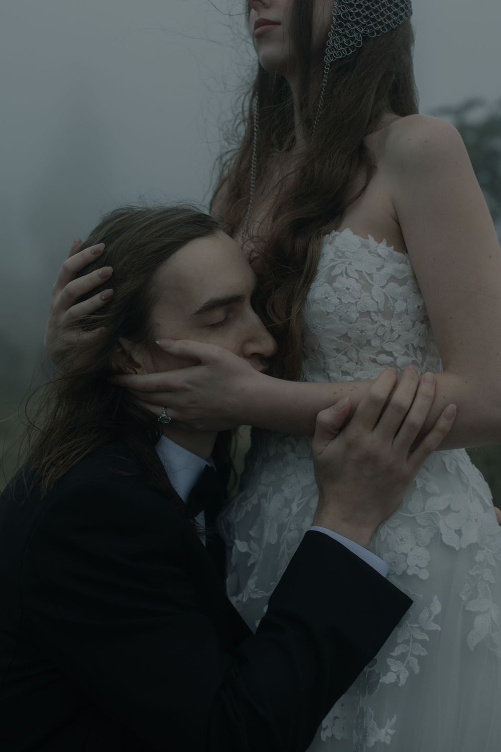 A groom in a black suit kneels before a bride in a white lace gown, sharing an intimate embrace in moody fog.