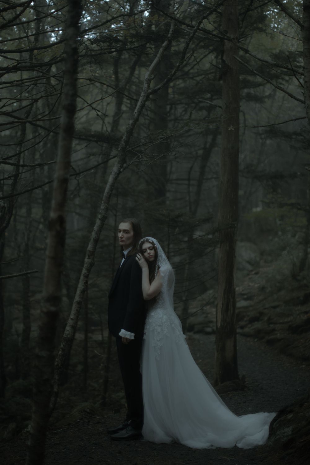 Bride and groom standing in a dark moody forest, bride wearing a white lace gown with veil, groom in black suit.