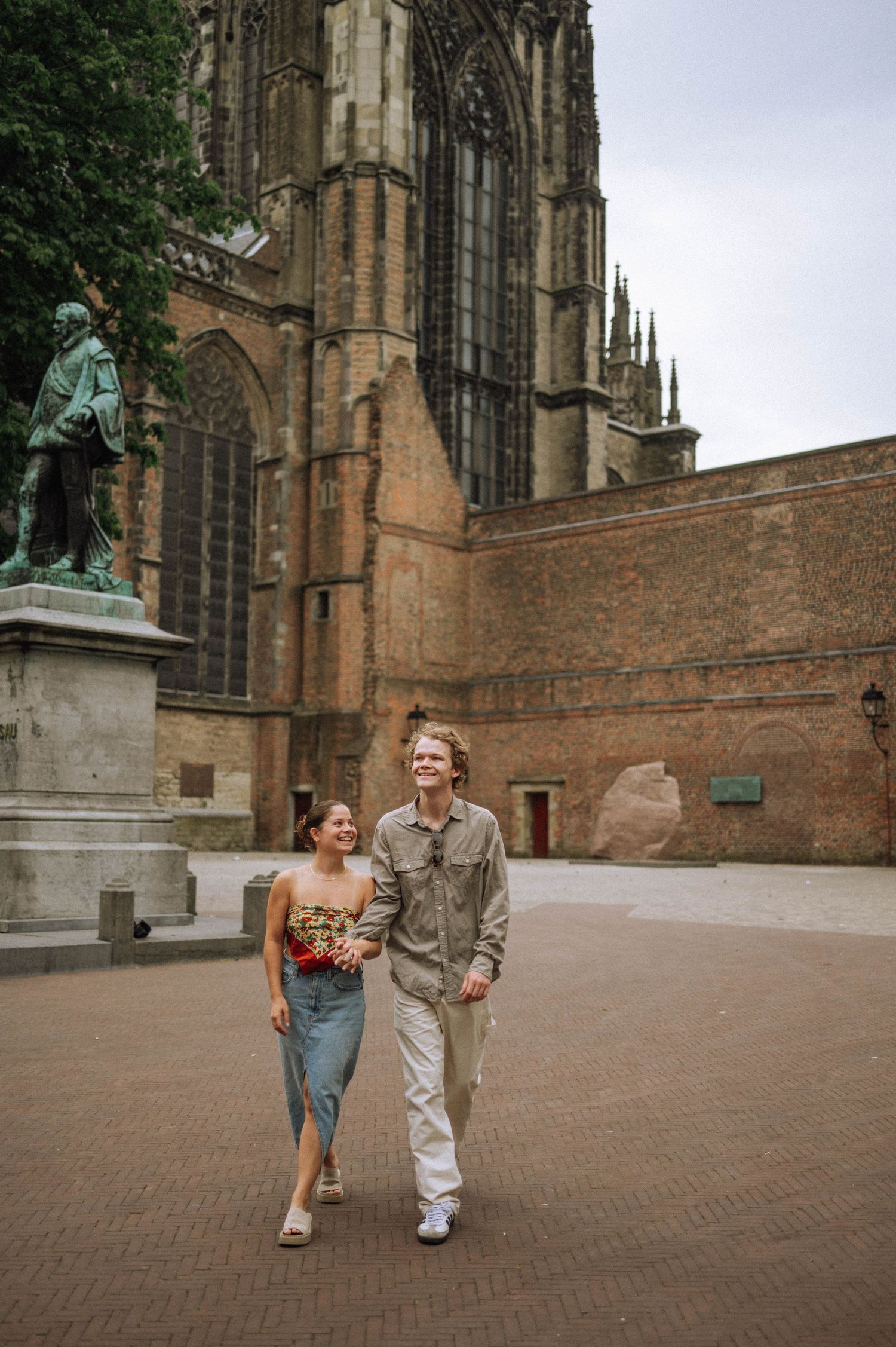 A joyful couple walking hand in hand through a square after a secret proposal, with the historic Utrecht Cathedral in the background.