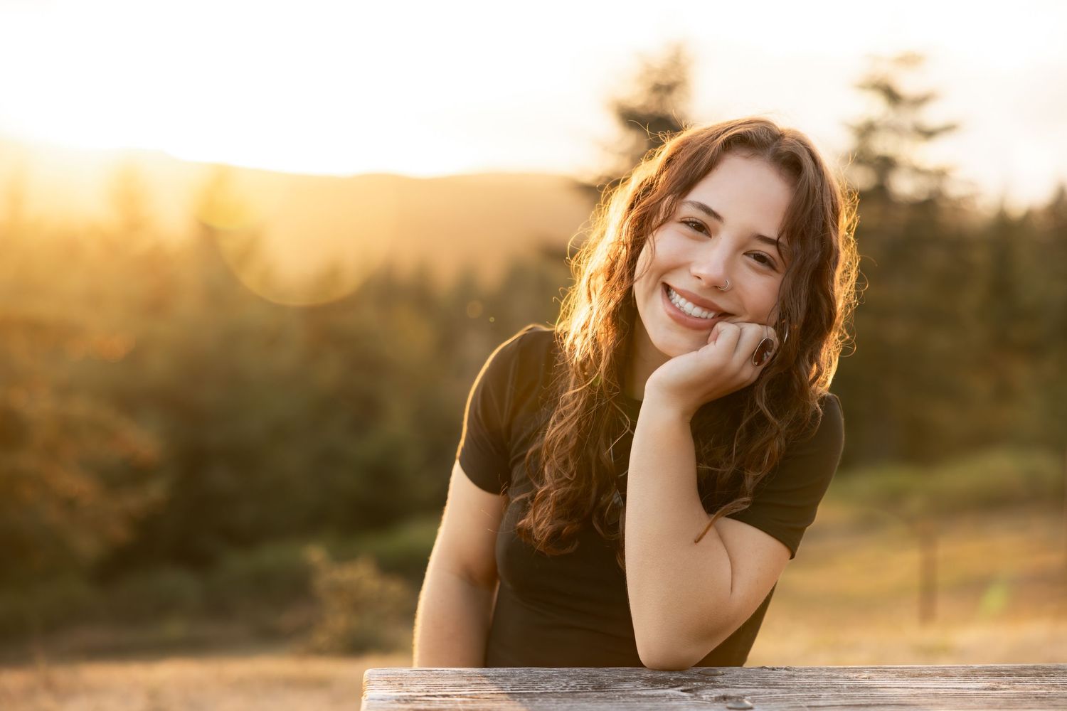 Person in black t-shirt smiling during golden hour sunset with forest in background.