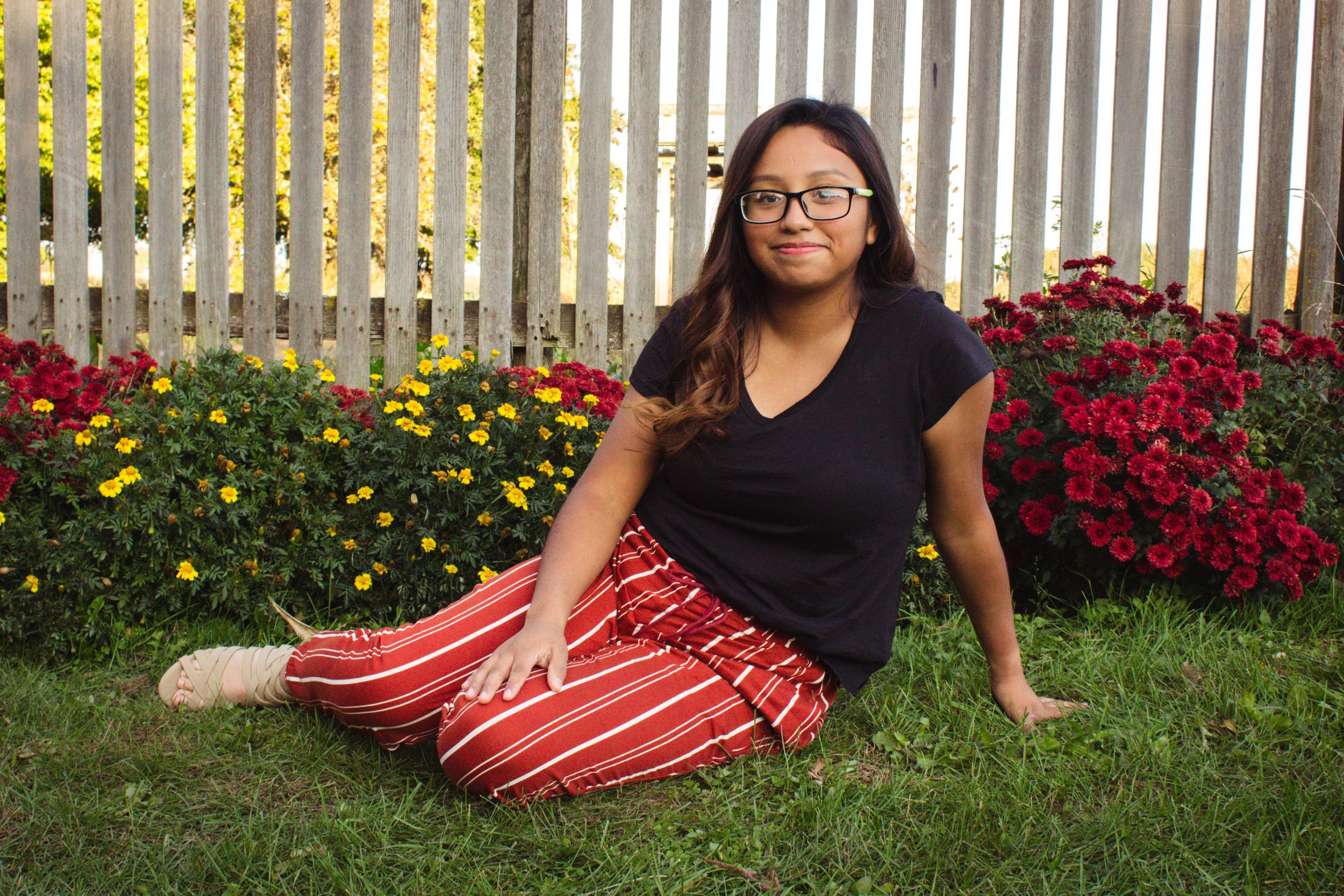 Someone relaxes on grass wearing a black top and striped red pants near colorful flower garden.