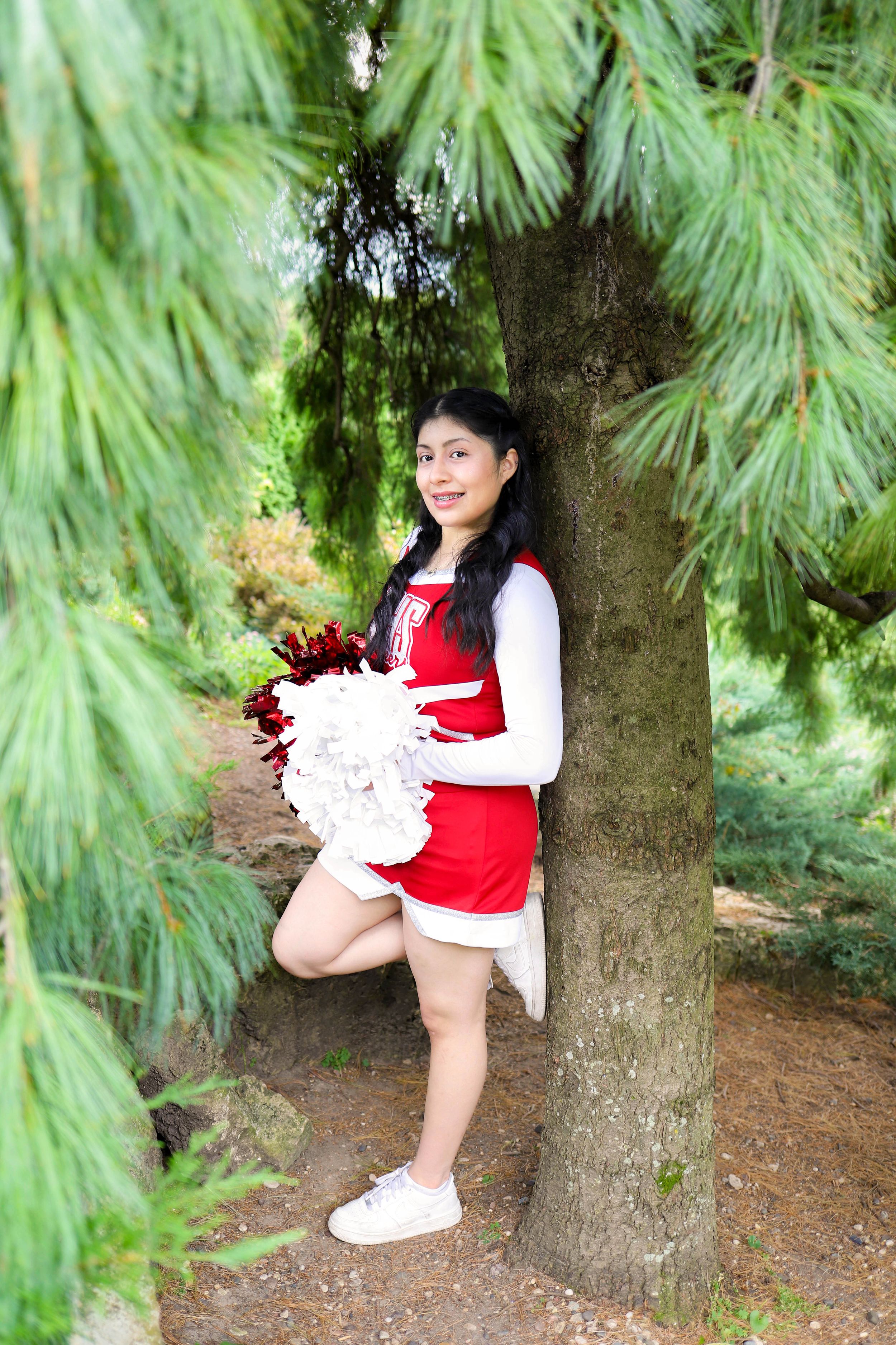 A cheerleader in red and white uniform poses next to a tree with pom poms.
