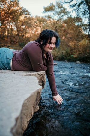 Person in brown shirt lying on rocky ledge reaching toward flowing stream water below.