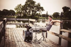 A vintage-toned photo of someone posing dramatically on a dock with a drum set.