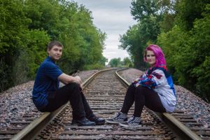 Two people sit on railroad tracks surrounded by green trees on both sides.