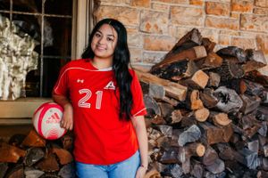 A soccer player poses with a ball in front of stacked firewood, wearing a red jersey numbered 21.