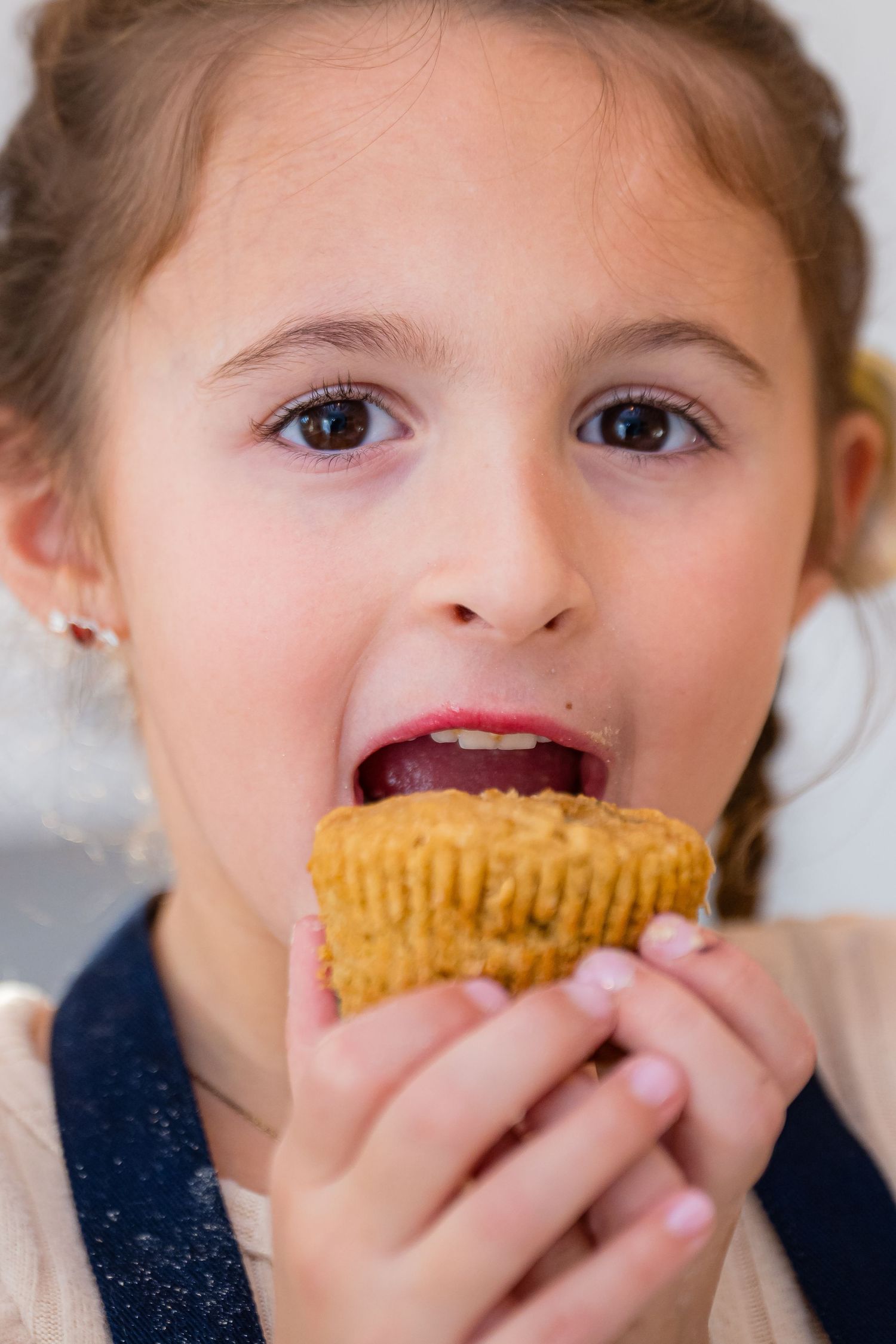 A young child takes a big bite of a golden cupcake while wearing a navy colored top.