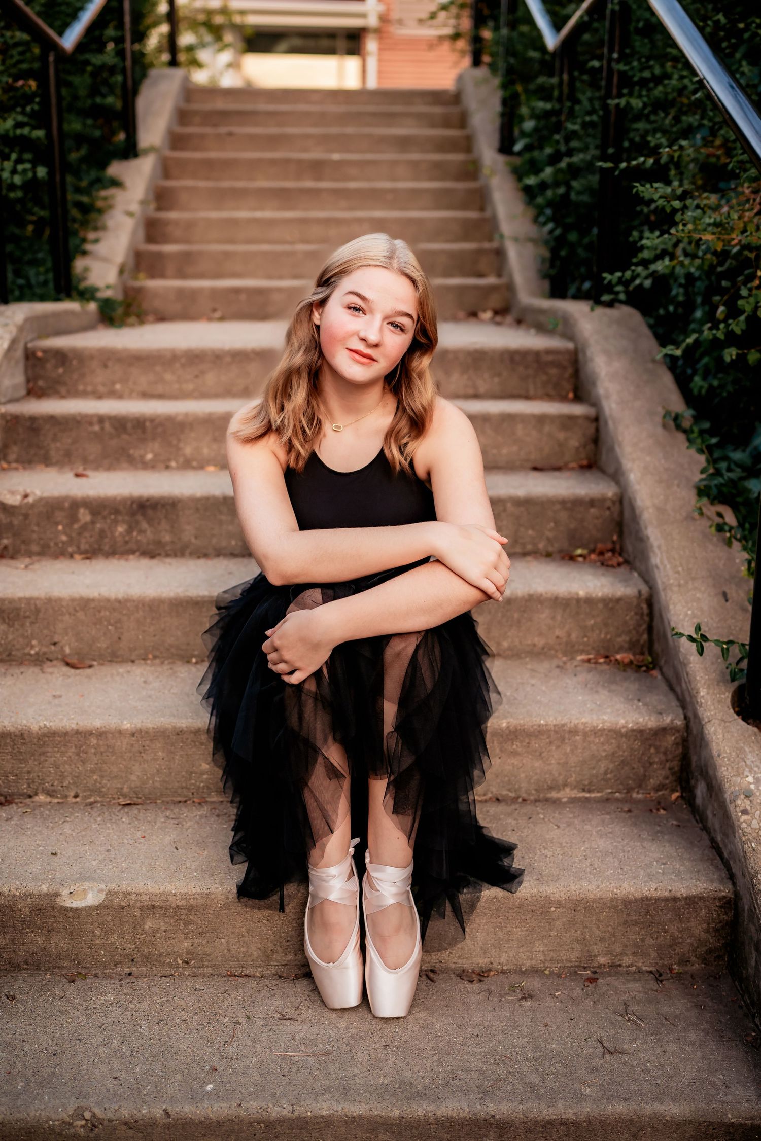 Ballet dancer in black tutu sits gracefully on concrete steps wearing pointe shoes.