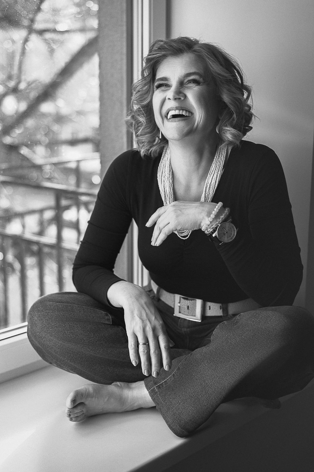 Black and white photo of a laughing woman sitting cross-legged on a windowsill wearing jeans and a dark top.