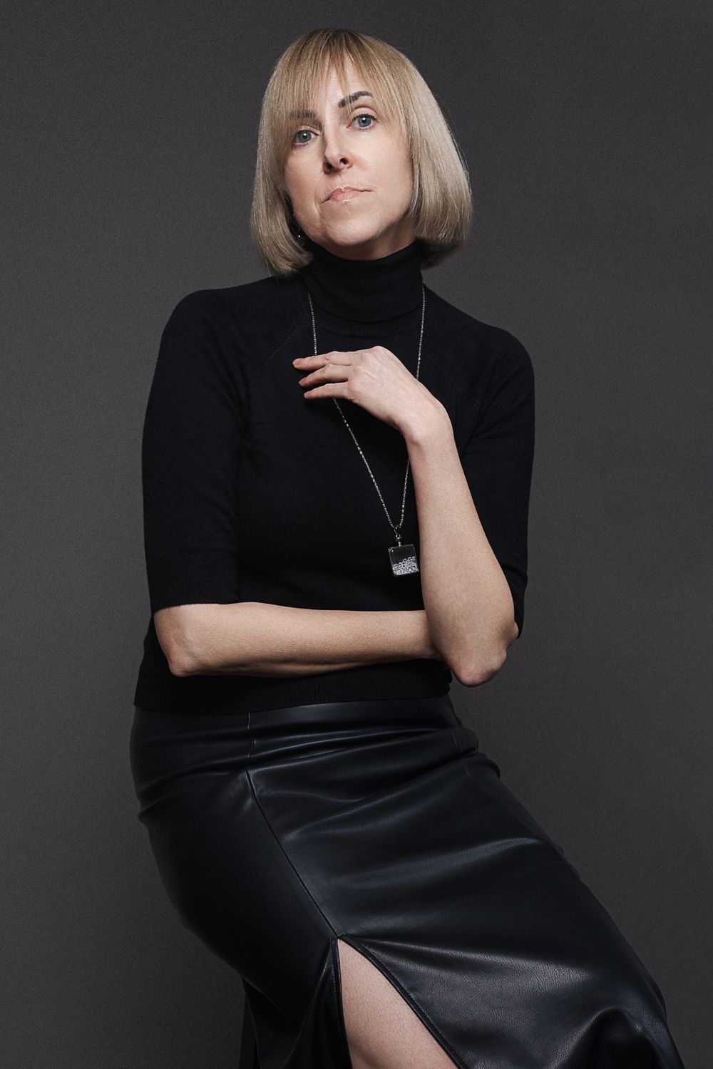 Professional studio portrait of a person in black turtleneck and leather skirt against dark background.