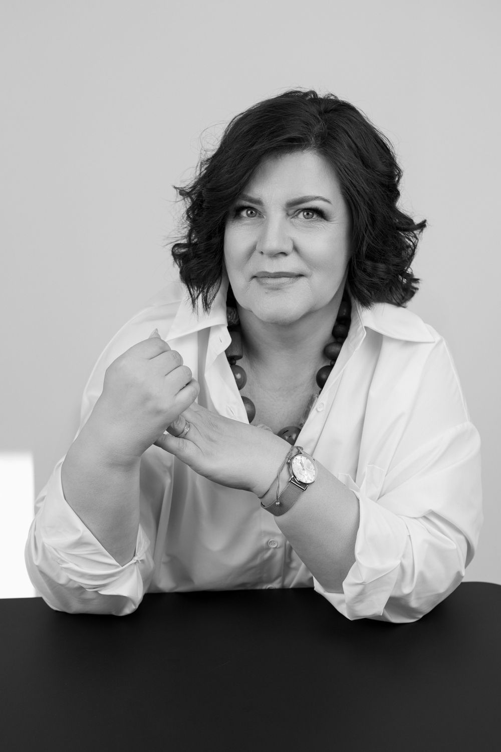 Black and white portrait of a confident woman in white shirt, sitting at desk with hands clasped, wearing a watch.