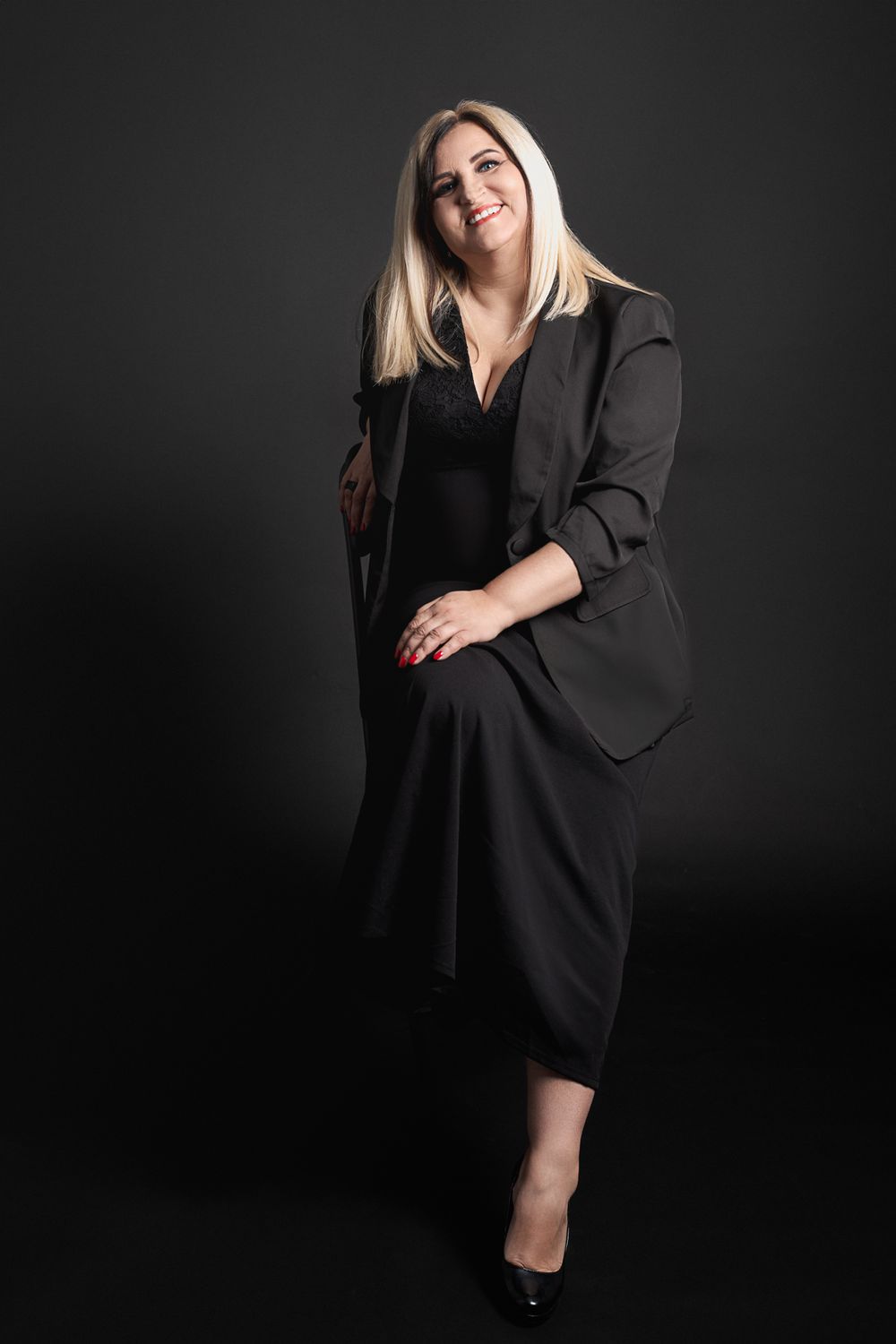 Blonde woman in black blazer and dress poses confidently against dark background in stylish studio portrait.