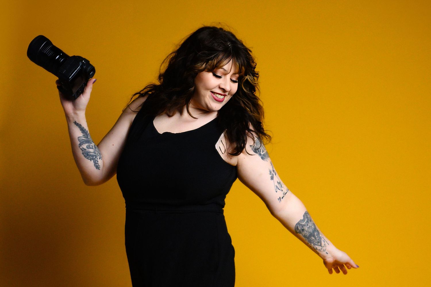 Joyful female photographer dancing and holding camera against a bright yellow background with tattoos.