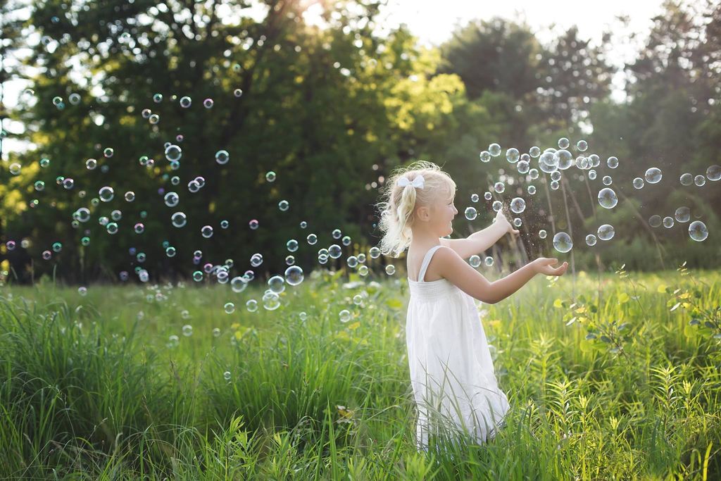 A child in a white dress plays with bubbles in a sunlit meadow during golden hour.