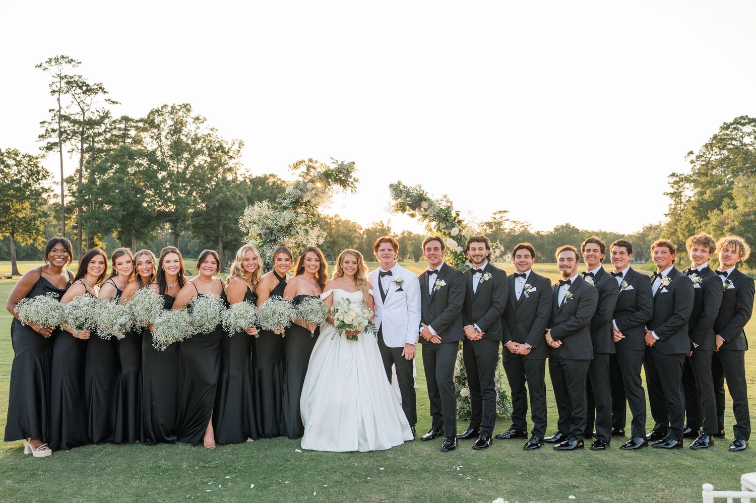 Wedding ceremony takes place outdoors with bridal party in black attire and pampas grass decor against natural landscape.