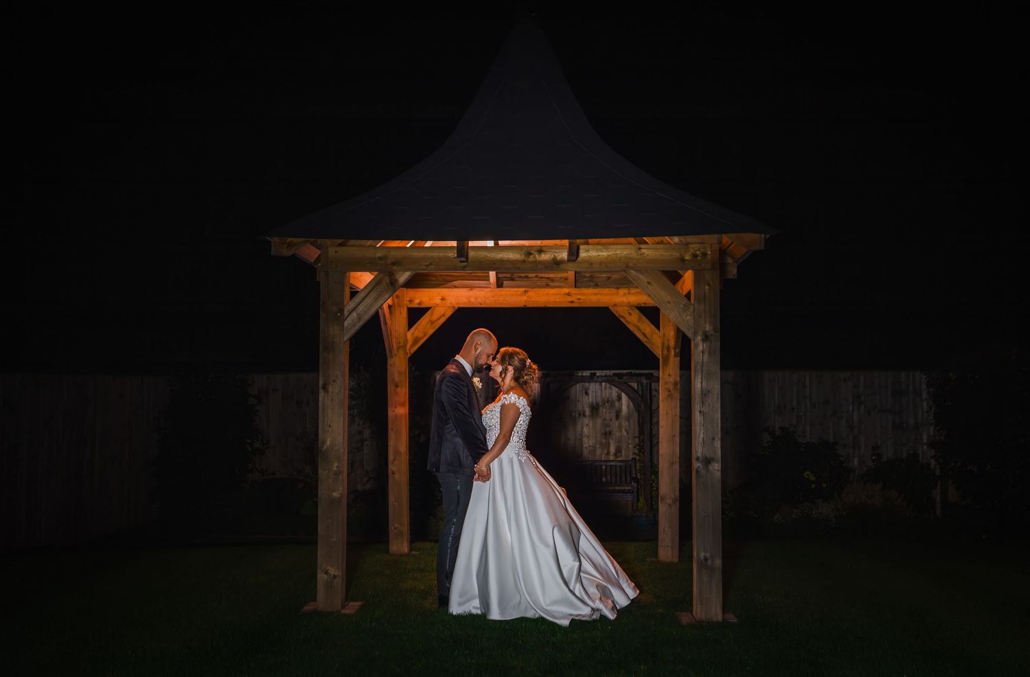 A dramatic nighttime wedding portrait under a wooden gazebo with orange lighting.
