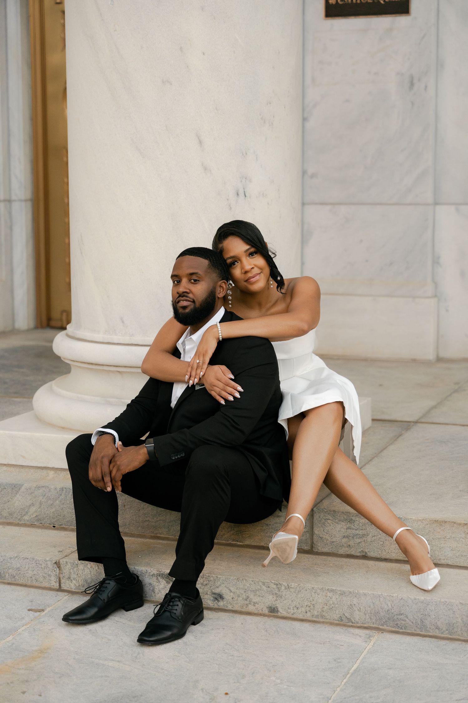 A couple poses together on marble steps, with her in a white dress leaning against him in a black suit.