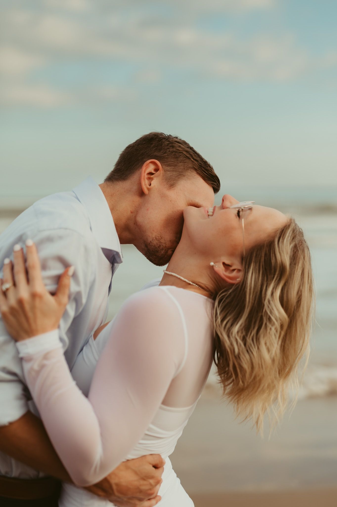 A couple shares a romantic kiss on the beach, with the woman in a white dress leaning back in her partner's arms.